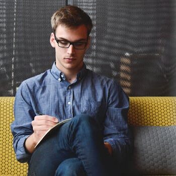 entrepreneur-startup-start-up-man-39866-39866 A young man in glasses writes in a notebook while sitting on a stylish couch indoors.